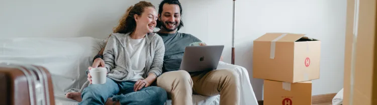 A couple sitting on a bed with a laptop and coffee, moving boxes stacked behind them.